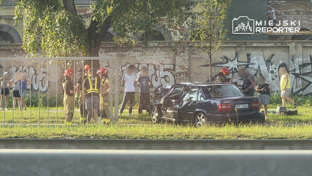 Grupa strażaków rozmawia obok uszkodzonego samochodu marki Volvo na tle graffiti i zarośli.