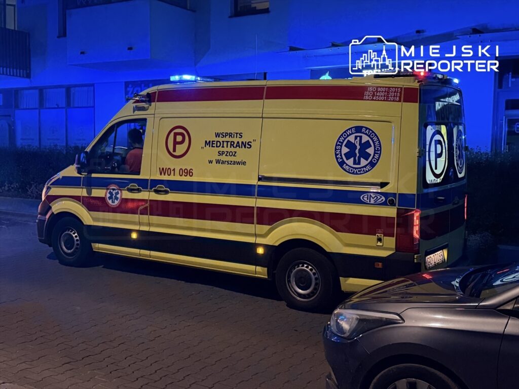 Karetka pogotowia z włączonymi sygnałami świetlnymi stoi na ulicy, obok zaparkowanego samochodu.