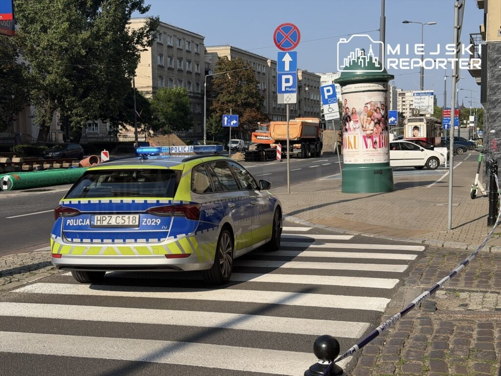 Policyjny radiowóz z niebieskimi światłami zatrzymany na przejściu dla pieszych na ruchliwej ulicy, obok słupka reklamowego.
