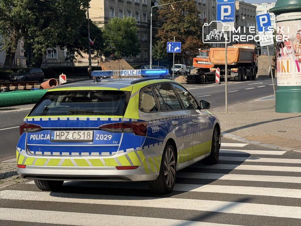 Policyjny radiowóz z niebieskimi światłami stoi na przejściu dla pieszych w miejskim otoczeniu, obok placu budowy.