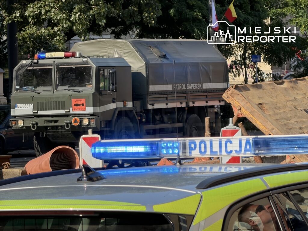 Patrol saperski z oznaczeniem „Uwaga, niewybuchy” stoi obok policyjnego radiowozu z włączonymi sygnałami świetlnymi.