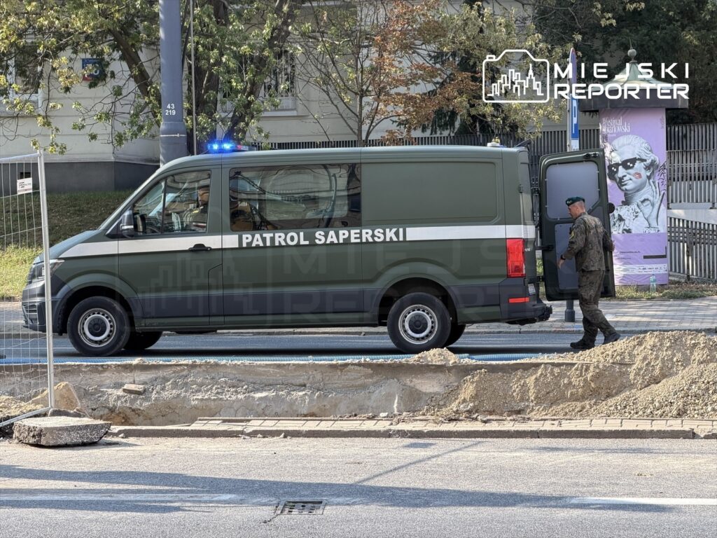 Żołnierz w mundurze z niebieskim beretem podchodzi do otwartych drzwi wojskowego pojazdu z napisem "PATROL SAPERSKI" obok wykopu na ulicy.