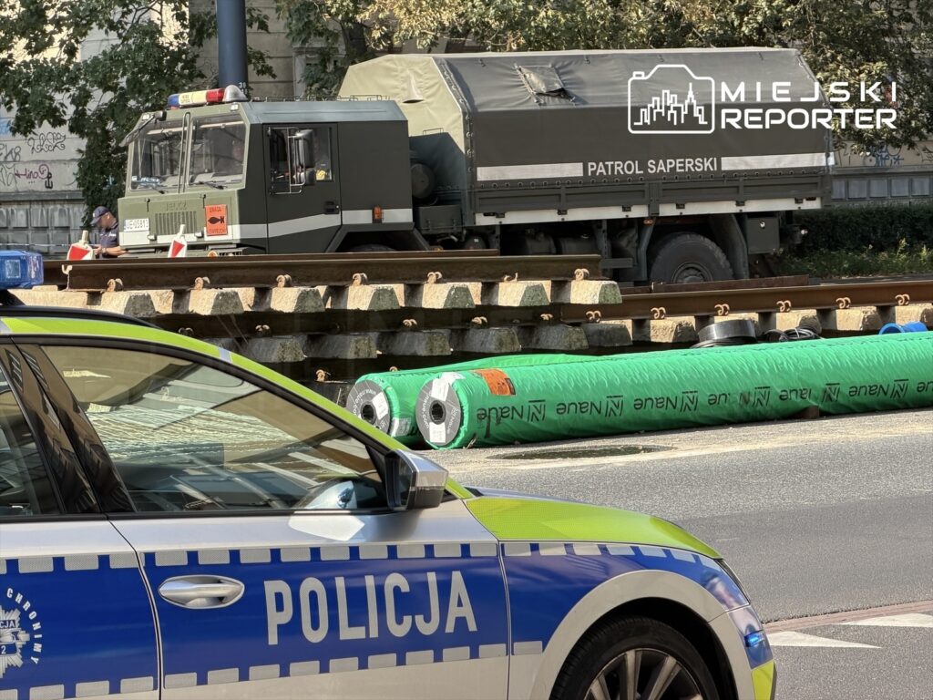 Patrol saperski z oznaczeniami na pojeździe stoi obok torów kolejowych, a w tle widoczny radiowóz policji.