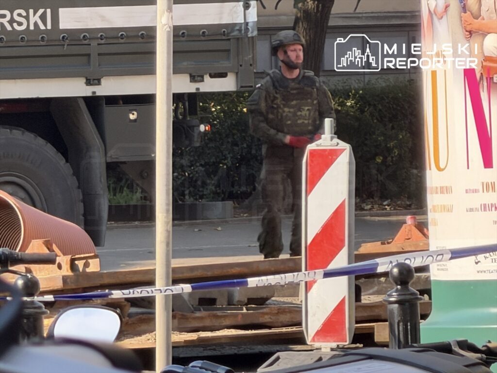 Żołnierz w mundurze stojący obok oznaczonego miejsca zabezpieczonego taśmą, w tle ciężarówka wojskowa.