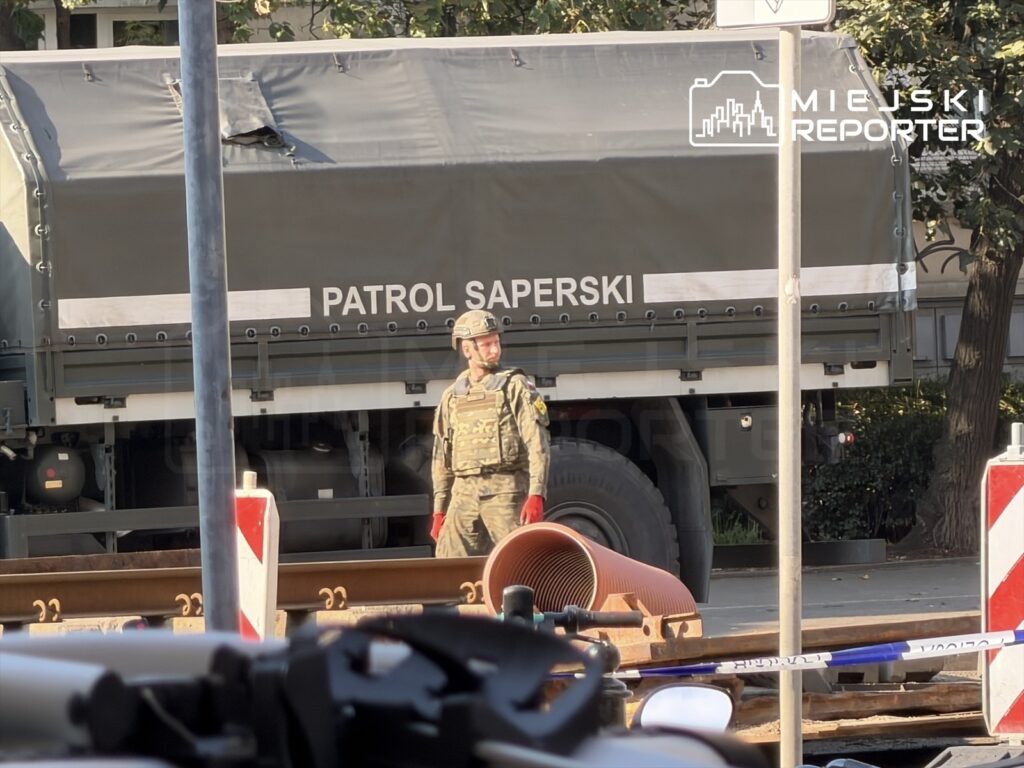 Żołnierz w mundurze stoi obok wojskowego pojazdu oznaczonego napisem „PATROL SAPERSKI” przy torach kolejowych.