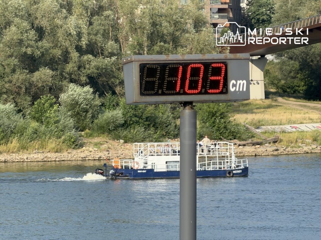 Tablica z cyfrowym pomiarem wskazującym 103 cm głębokości Wisły, obok płynąca łódź na rzece, w tle roślinność.