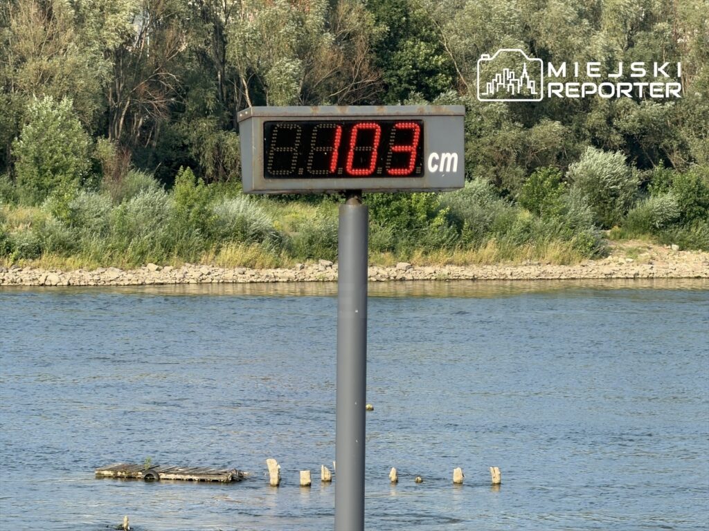 Wyświetlacz z oznaczeniem 0,03 cm wskazuje niski poziom wody w rzece Wisła przy Bulwarach. W tle widać zieleń i brzeg rzeki.