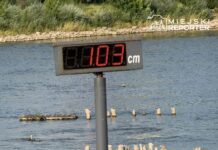 Wyświetlacz z cyfrowym wskazaniem poziomu wody na rzece, pokazujący 103 cm, w tle woda Wisły i kamienie brzegowe.