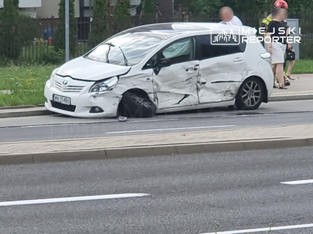 Uszkodzony biały samochód osobowy stoi na poboczu drogi, obok grupy osób w mundurach strażackich, Mokotów.