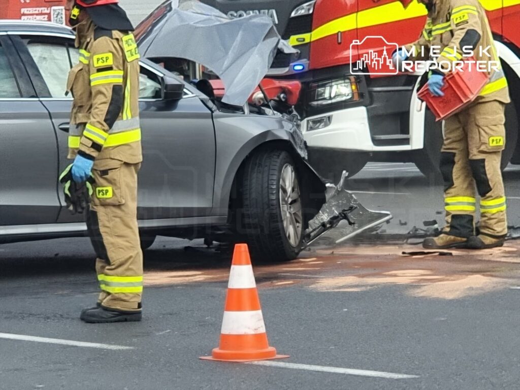 Strażacy w mundurach interweniują przy uszkodzonym samochodzie po wypadku na ulicy, obok pomarańczowego pachołka.