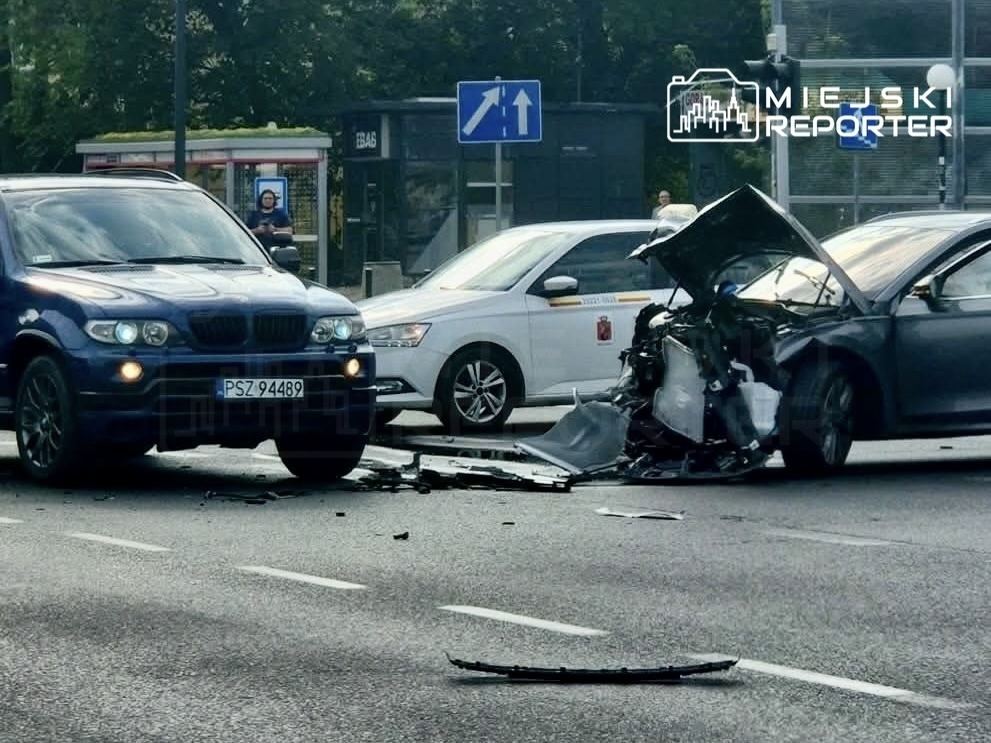 Dwa uszkodzone samochody na ulicy, z widocznymi zniszczeniami przodu jednego z nich, w tle przechodnie i znaki drogowe na Mokotowie.