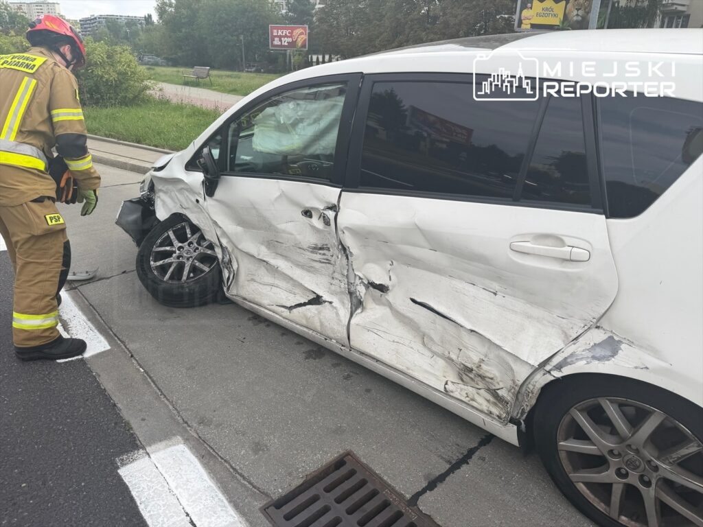Strażak w mundurze bada uszkodzone auto po wypadku drogowym na Mokotowie, widoczne zniszczenia karoserii.