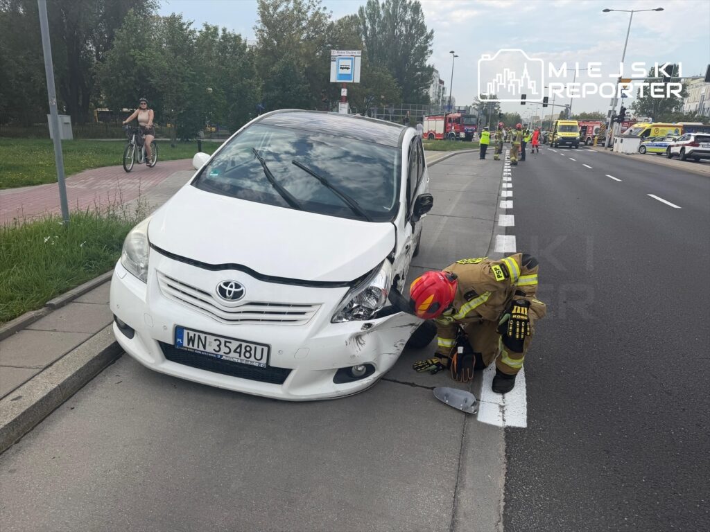 Strażak sprawdzający uszkodzenia białego samochodu osobowego na Mokotowie po wypadku drogowym. W tle inne pojazdy ratunkowe.