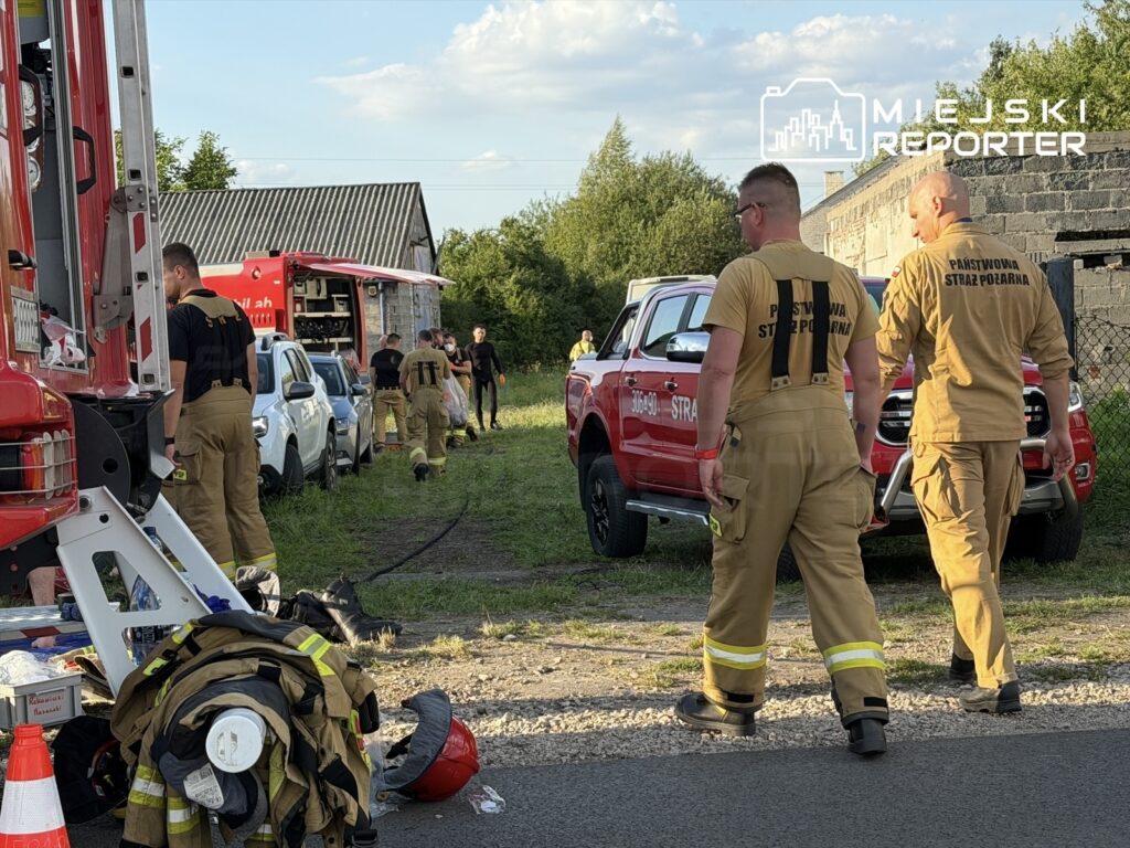 Grupa strażaków w mundurach pracuje przy wozach strażackich w terenie wiejskim, obok budynku.