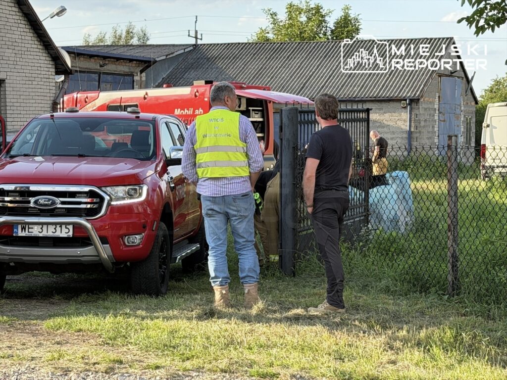 Mężczyzna w kamizelce odblaskowej rozmawia z innym mężczyzną obok czerwonego samochodu strażackiego na terenie wiejskim.