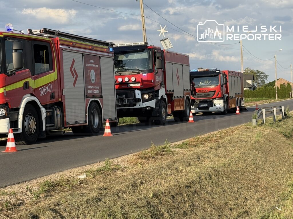 Dwa samochody strażackie zaparkowane na drodze, otoczone pachołkami, w tle pola i zabudowania.