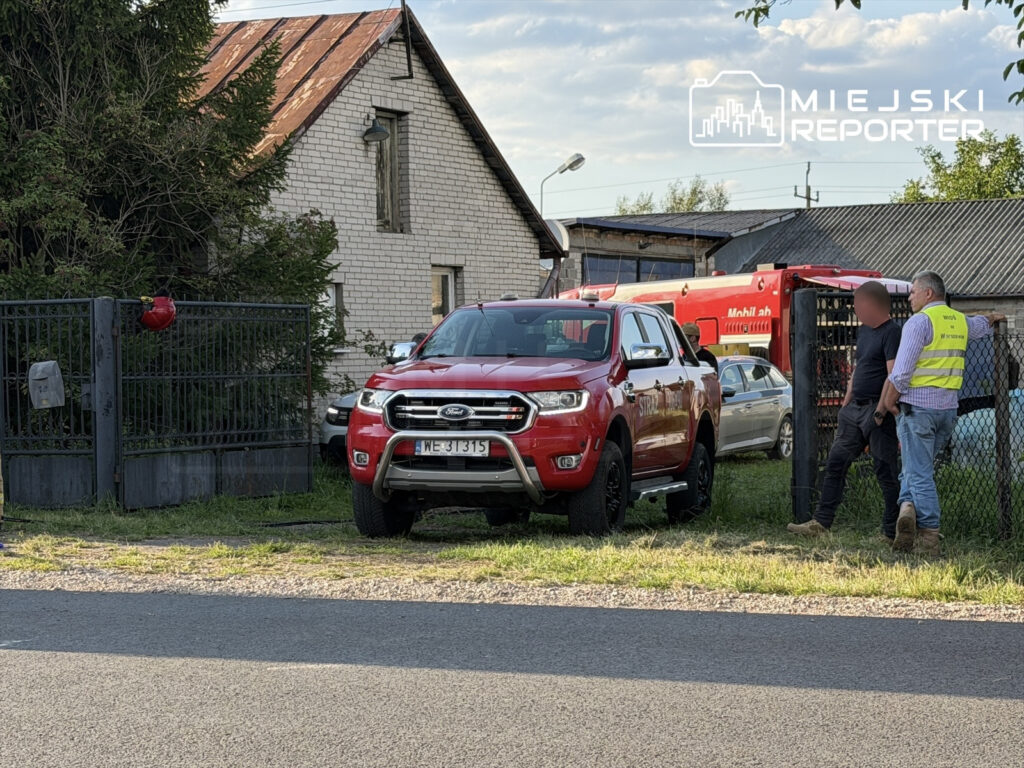 Czerwony samochód terenowy stoi przed bramą, obok dwóch mężczyzn rozmawiających w pobliżu budynku.