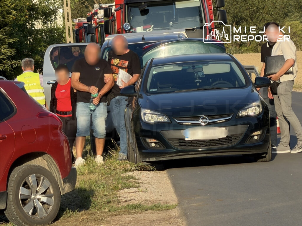 Grupa osób w różnych strojach stoi obok czarnego samochodu, a w tle widoczne są wozy strażackie.