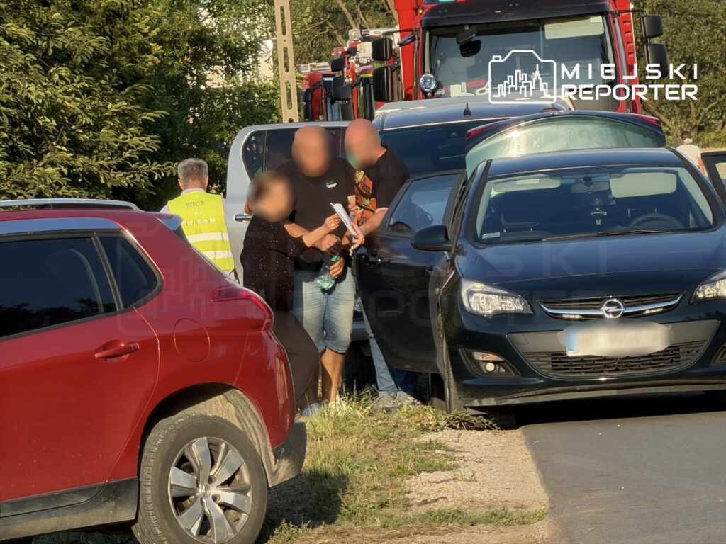 Mężczyzna w czarnej koszulce rozmawia z osobą w żółtej kamizelce, obok zaparkowane samochody i straż pożarna.