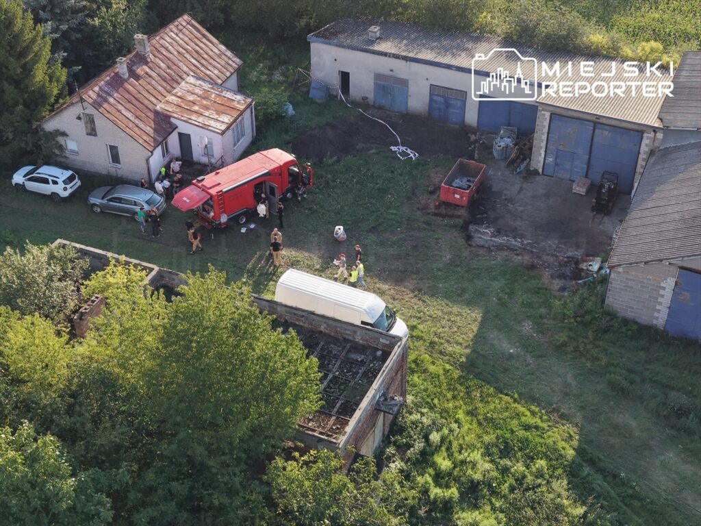 Grupa osób w mundurach rozmawia obok czerwonego wozu strażackiego na terenie wiejskim z budynkami gospodarczymi.