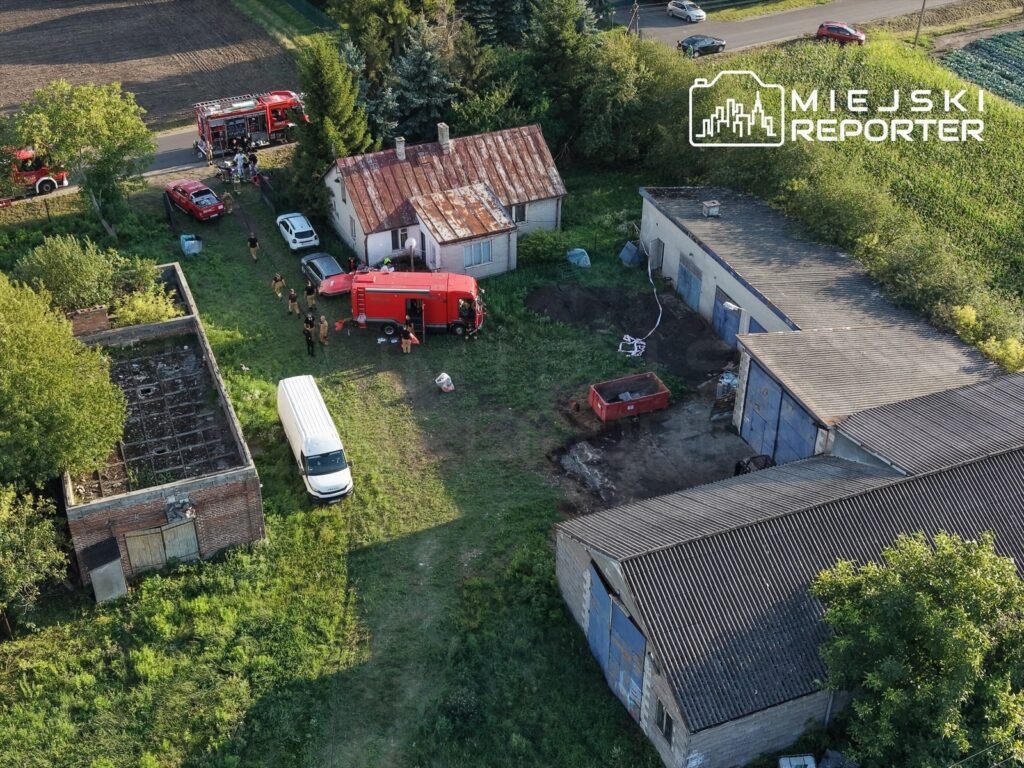 Grupa strażaków pracuje przy czerwonym wozie strażackim obok zrujnowanego budynku w terenie wiejskim.