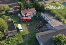 Grupa strażaków pracuje przy czerwonym wozie strażackim obok zrujnowanego budynku w terenie wiejskim.