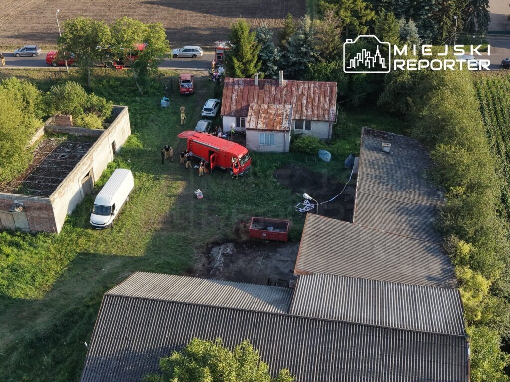 Grupa strażaków pracuje przy czerwonym wozie strażackim na terenie zarośniętej działki z opuszczonymi budynkami.