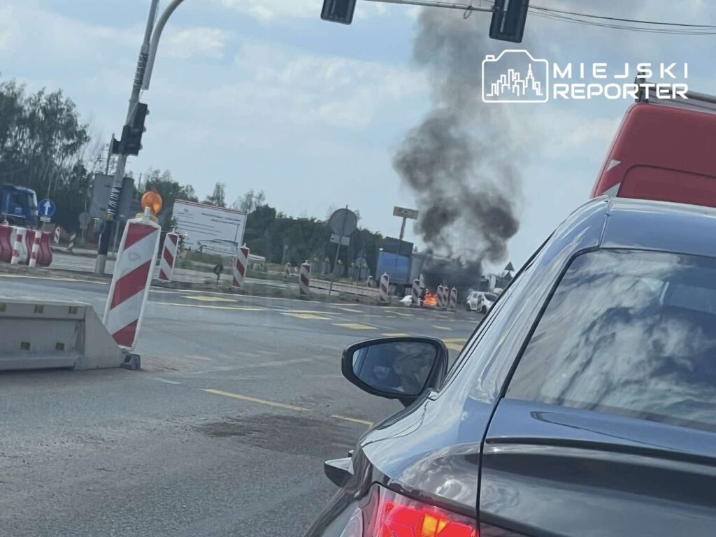 Auto płonie na jezdni, z dymem unoszącym się w powietrze, w tle znaki drogowe i barierki budowlane.
