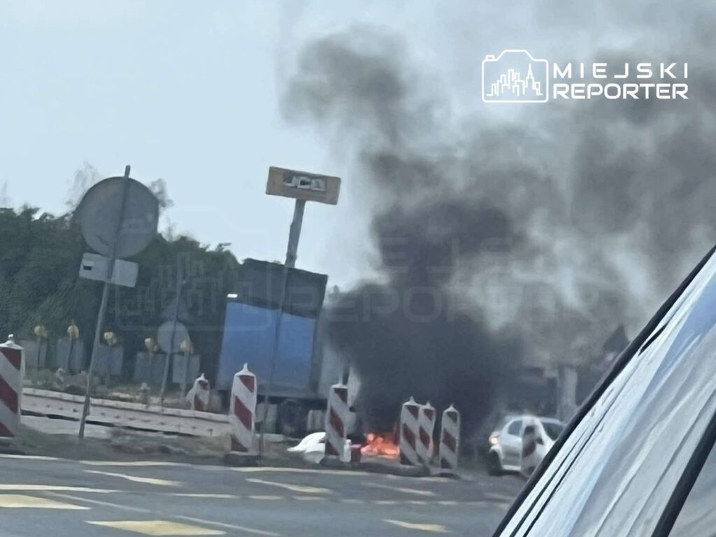 Auto płonie na poboczu drogi, z dymem unoszącym się w powietrzu, obok widoczna ciężarówka i znaki drogowe.