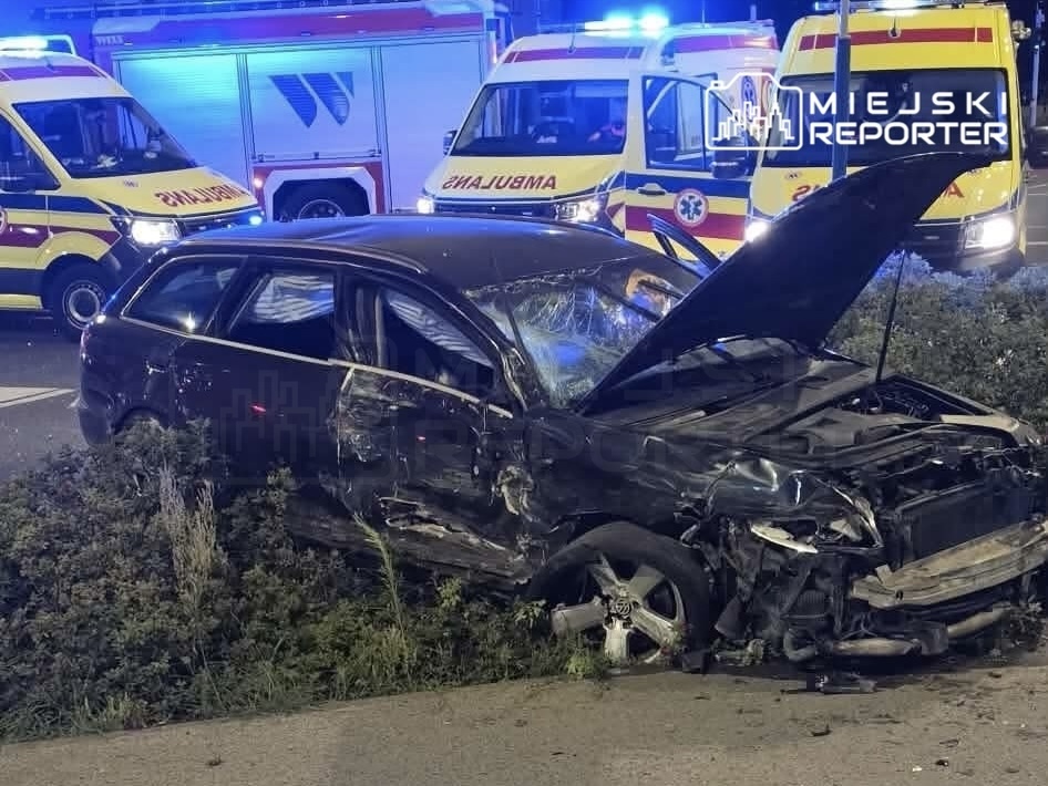 Czarny samochód osobowy z uszkodzoną karoserią stoi na trawie obok drogi, w tle widoczne karetki pogotowia.