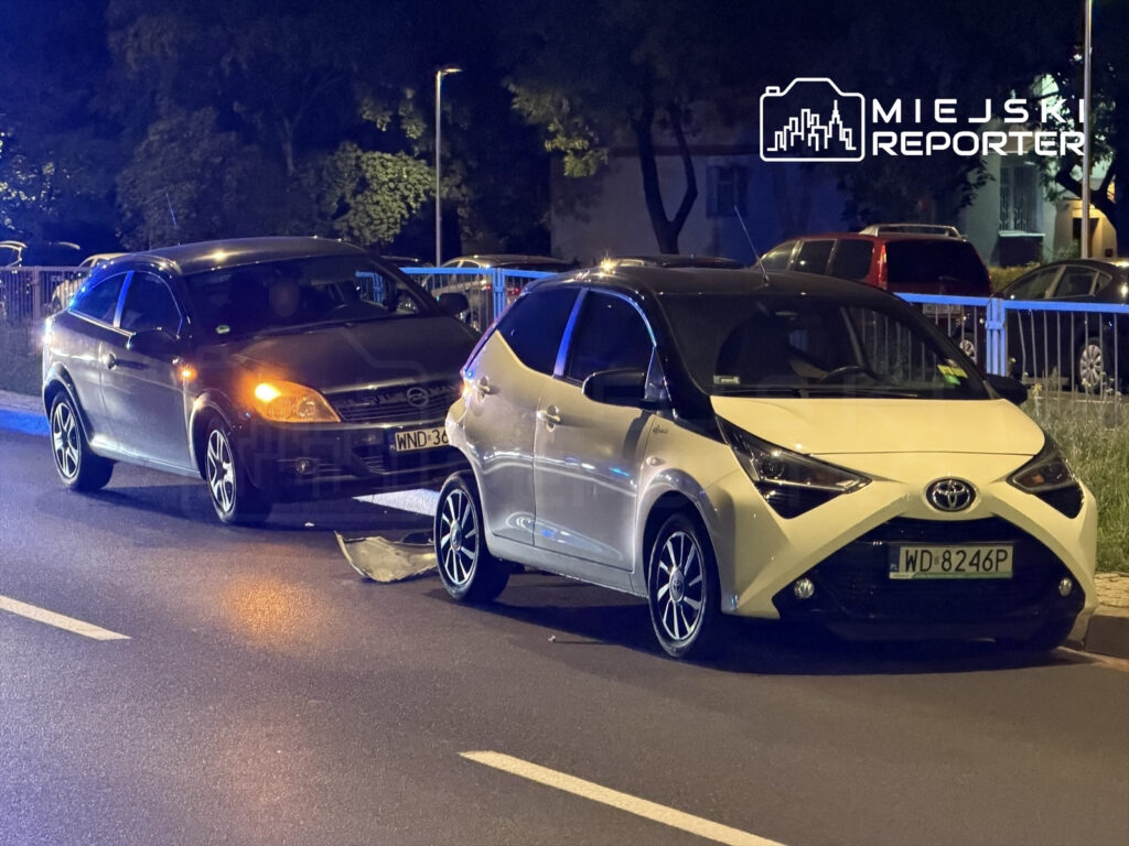 Dwa samochody osobowe, czarny i biały, stoją na drodze po kolizji, z uszkodzeniami widocznymi na karoserii.