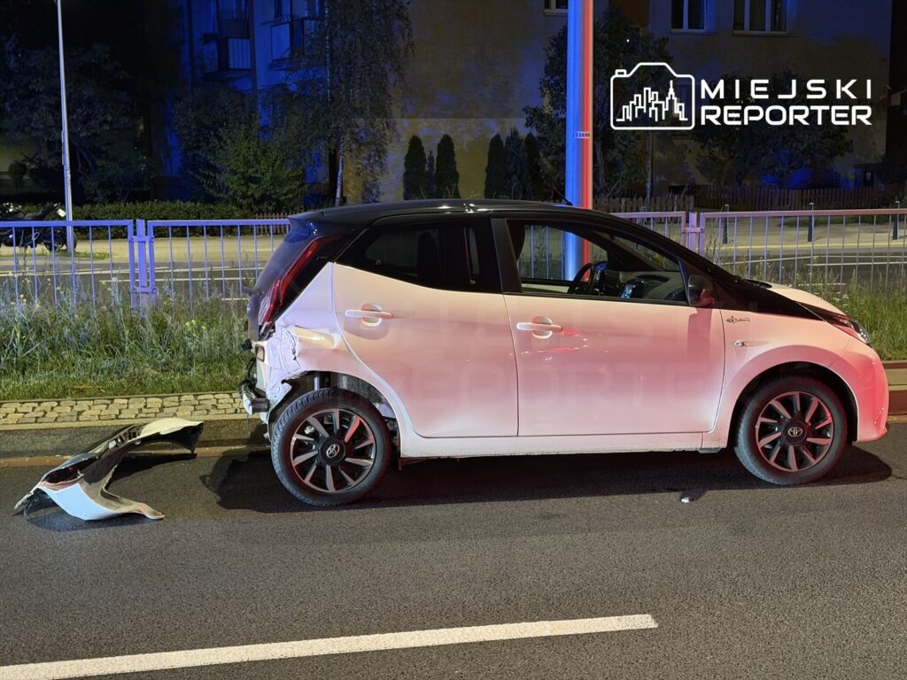 Czarny samochód osobowy z uszkodzoną tylną częścią stoi na jezdni, obok leży odłamany zderzak. W tle widoczne oświetlenie nocne.