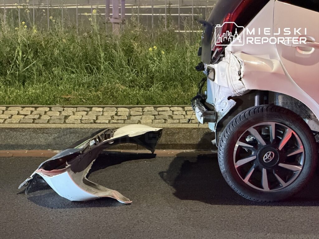 Uszkodzony tył białego samochodu osobowego z oderwaną częścią karoserii leżącą na jezdni. W tle widać trawnik i chodnik.