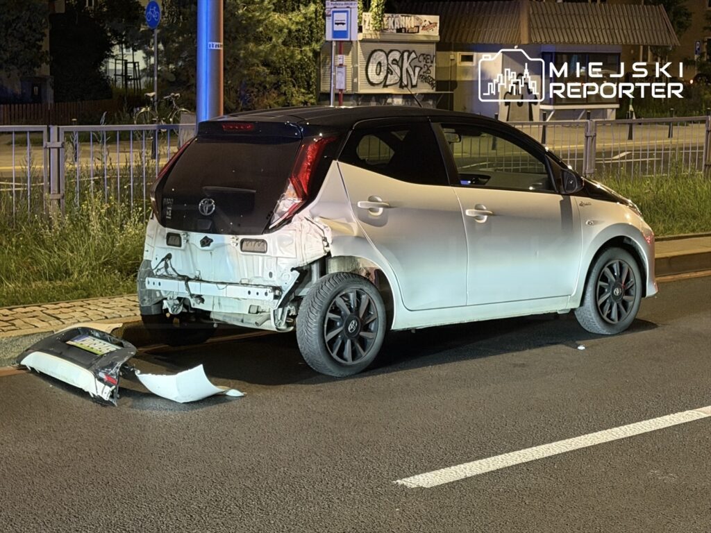 Uszkodzony biały samochód zaparkowany na poboczu, z odłączonym zderzakiem leżącym obok, w nocy na miejskiej ulicy.