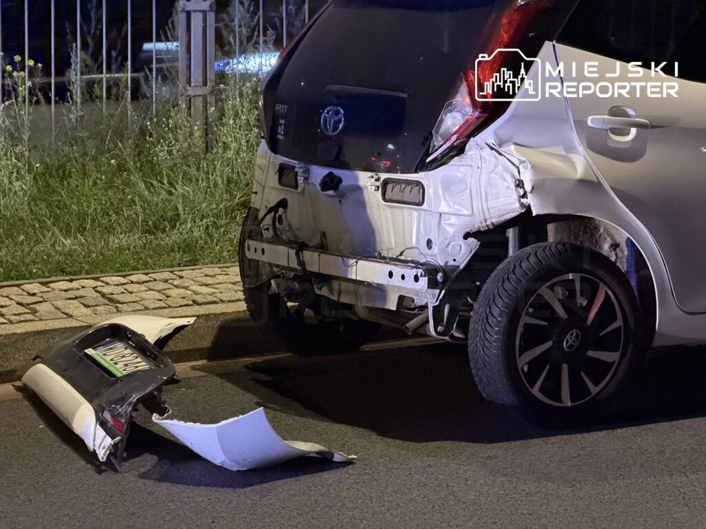 Zniszczony tył białego samochodu osobowego leży na drodze, obok znajduje się oderwana część zderzaka.