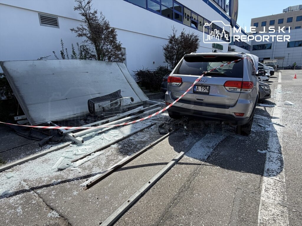 Jeep z uszkodzoną tylną częścią stoi na parkingu obok rozbitej wiaty i wózka sklepowego, wokół szkło.