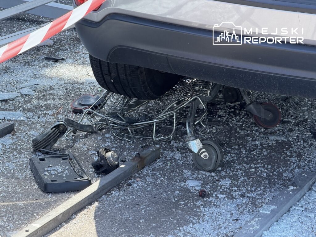 Samochód osobowy stoi na zgniecionej wózku sklepowym, otoczony odłamkami szkła na parkingu.