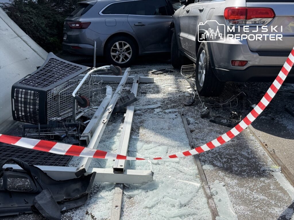Zniszczone wózki sklepowe i rozbite szkło leżą na ziemi obok uszkodzonego samochodu w miejskim otoczeniu.
