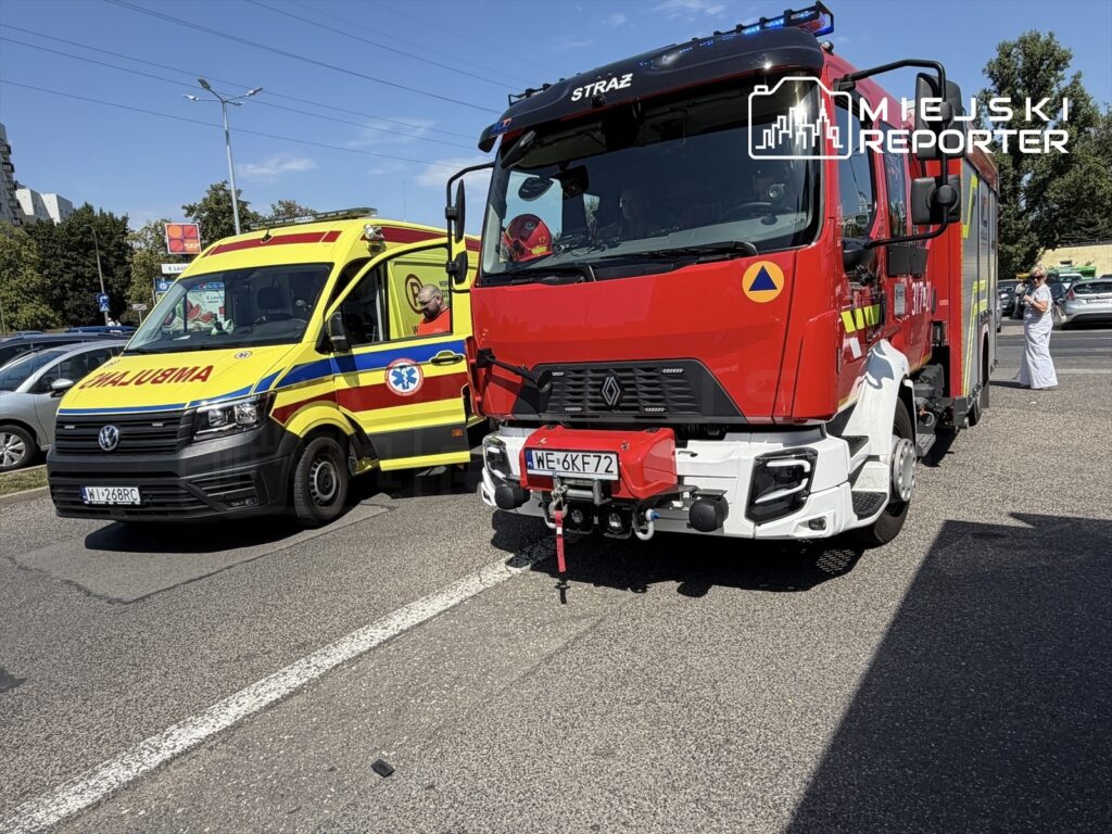 Ambulans i wóz strażacki stoją obok siebie na ruchliwej ulicy, w tle widoczne zaparkowane samochody.