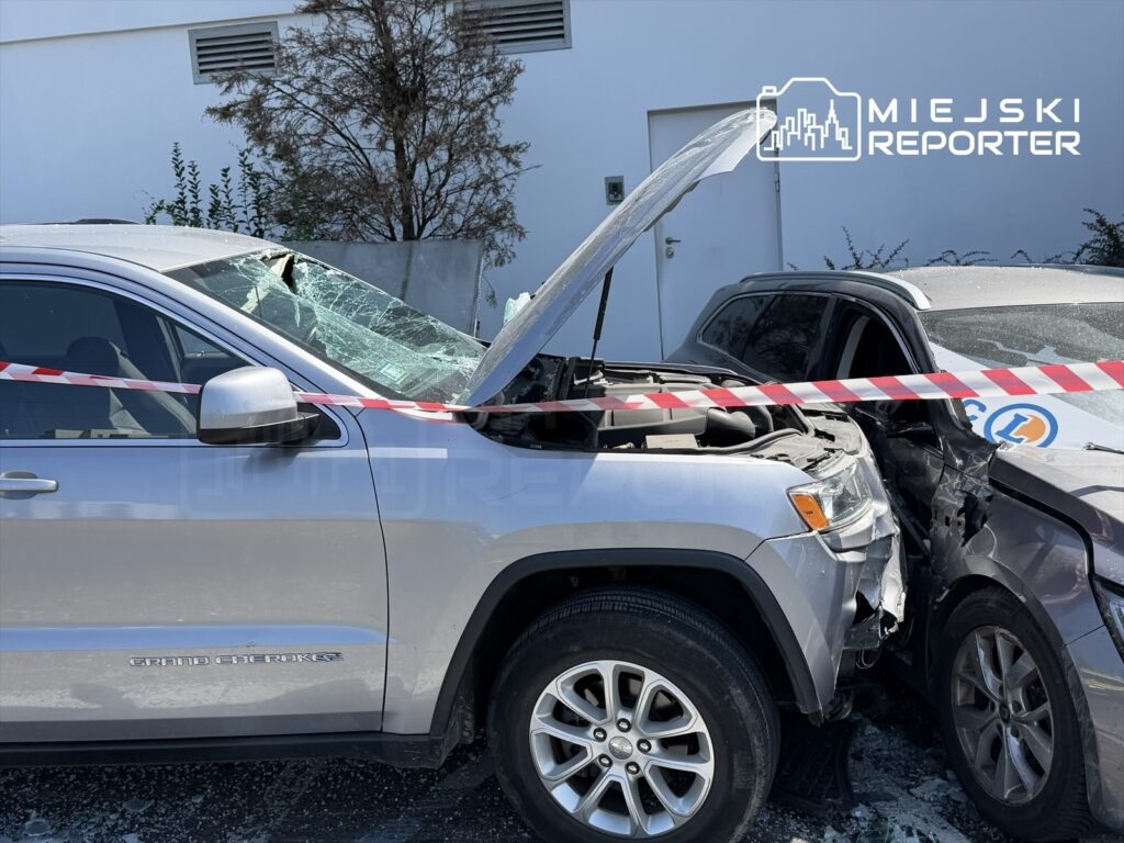 Jeep z otwartą maską stojący obok uszkodzonego samochodu, otoczony taśmą ostrzegawczą, z rozbitą szybą.