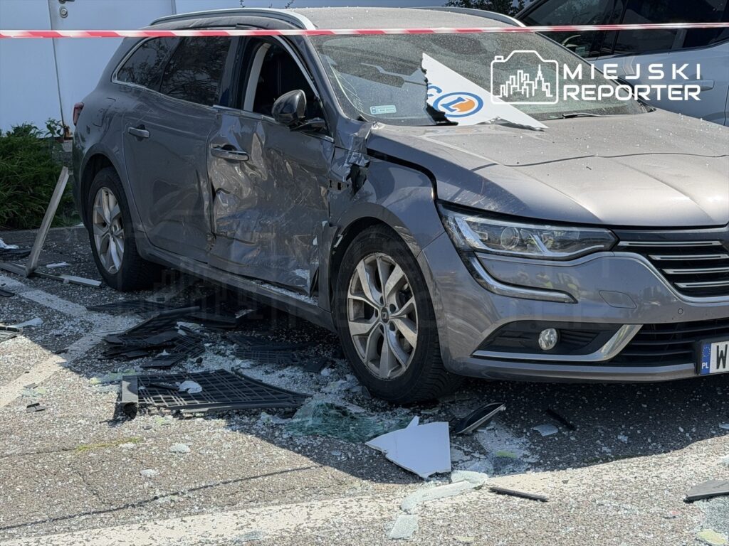 Uszkodzony samochód osobowy z wgniecioną stroną stoi na parkingu, wokół rozbite szkło i fragmenty wózków sklepowych.