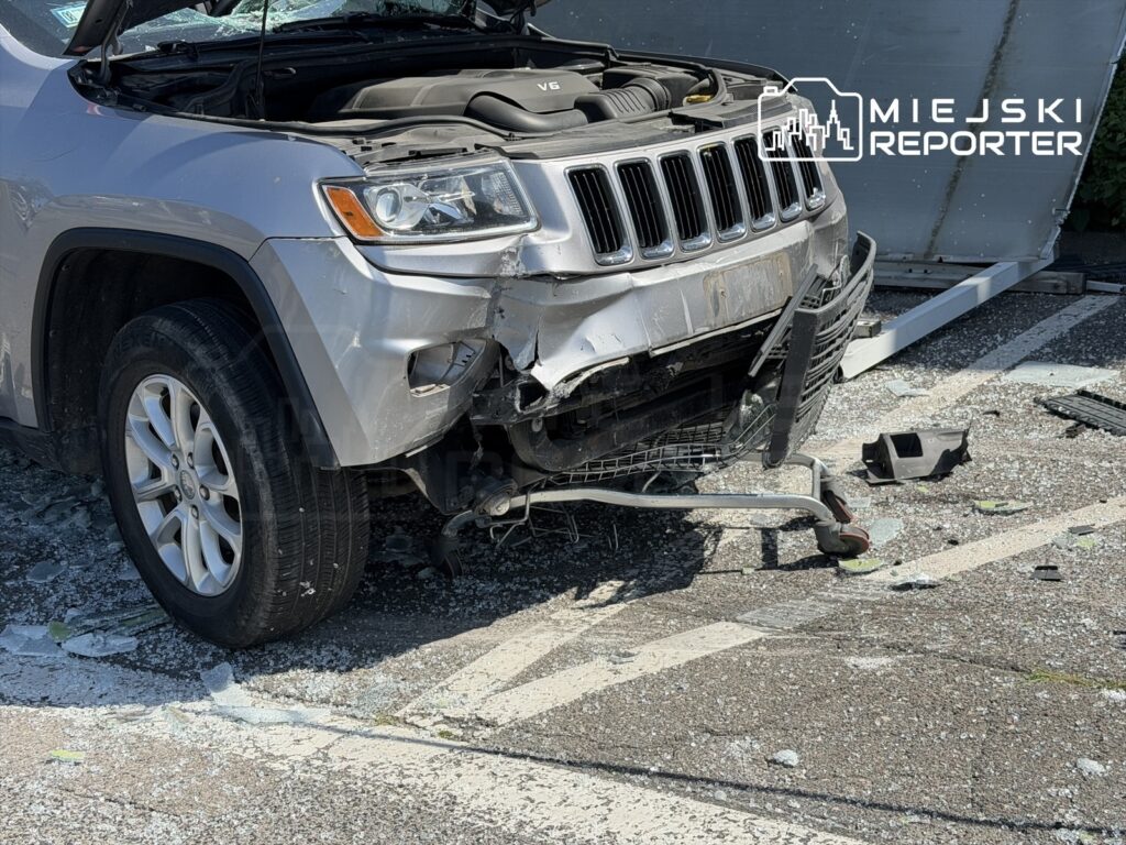 Uszkodzony srebrny Jeep z zniszczonym przodem stoi na parkingu, otoczony fragmentami szkła i odłamkami.