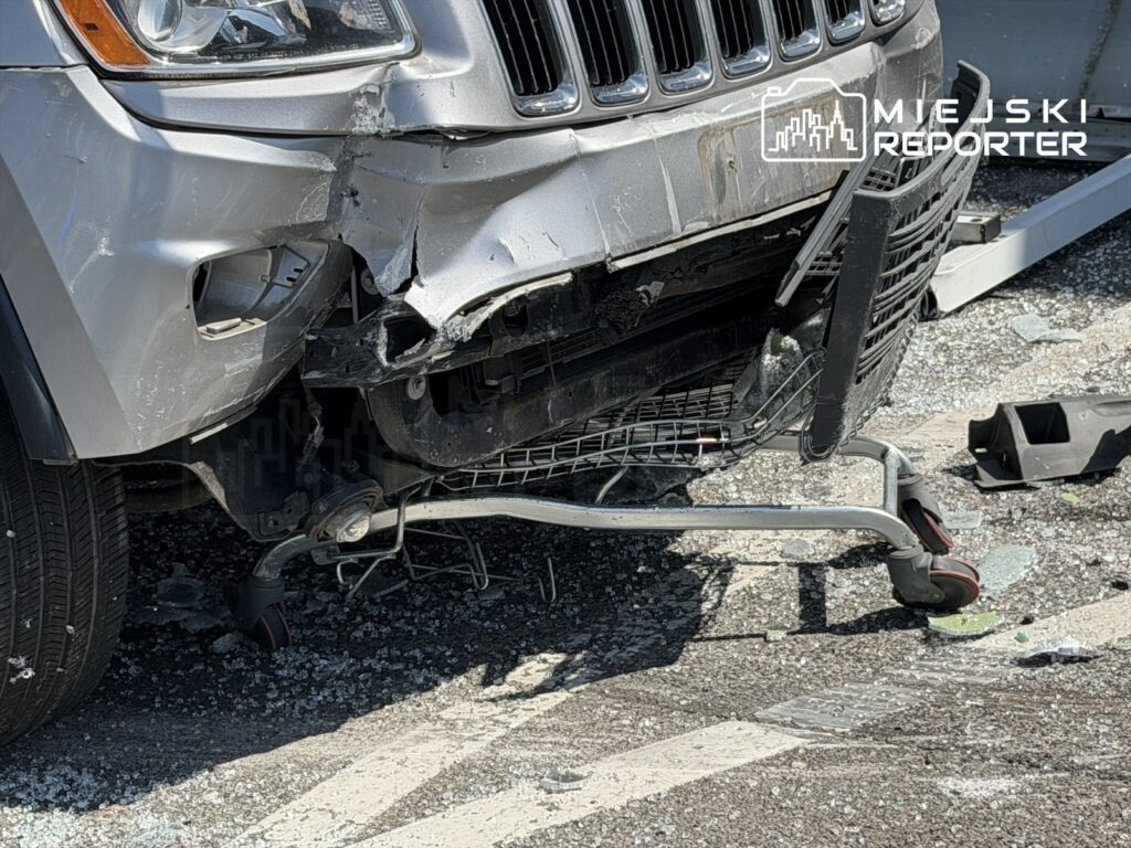 Uszkodzony przód srebrnego SUV-a z wygiętą maską, obok wózek sklepowy i rozbite szkło na ziemi.