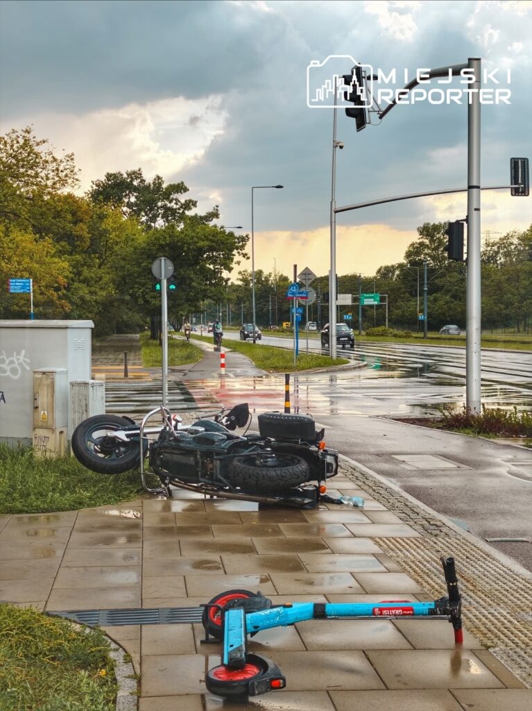 Przewrócony motocykl i niebieska hulajnoga leżą na chodniku obok drogi, w tle widoczne samochody i sygnalizacja świetlna.