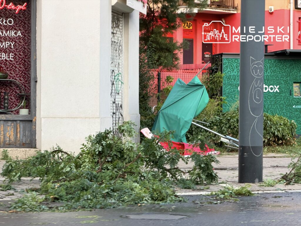 Powalone gałęzie i zniszczony parasol leżą na chodniku po burzy, obok zamkniętego lokalu.