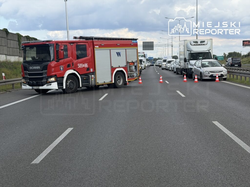 Wóz strażacki z włączonymi sygnałami blokuje pas ruchu na autostradzie, a samochody stoją w zatorze.