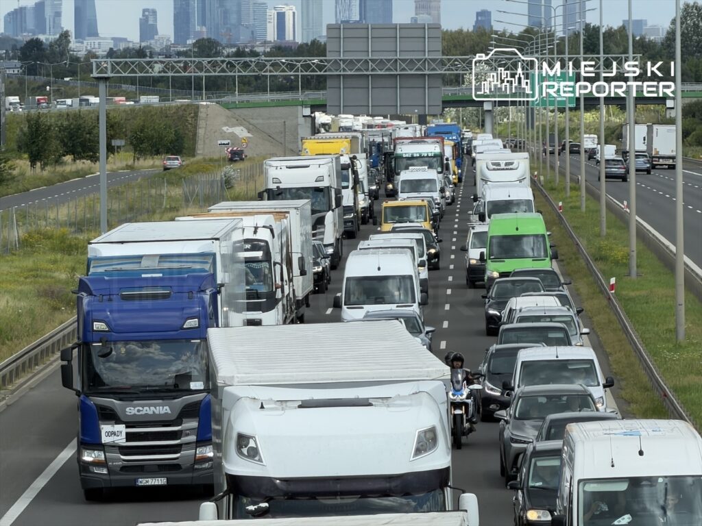 Zatłoczona autostrada z wieloma samochodami osobowymi i ciężarówkami, w tle wieżowce miasta.