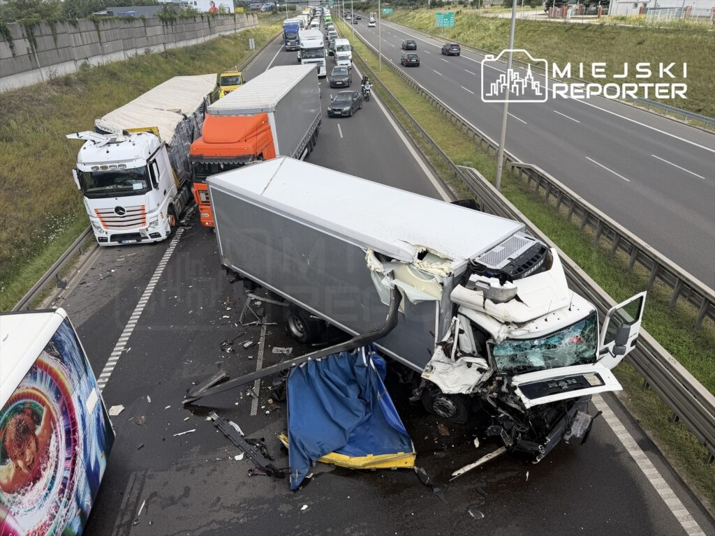 Zderzenie dwóch ciężarówek na autostradzie, z jedną z nich uszkodzoną i leżącą na boku. W tle widoczny ruch drogowy.