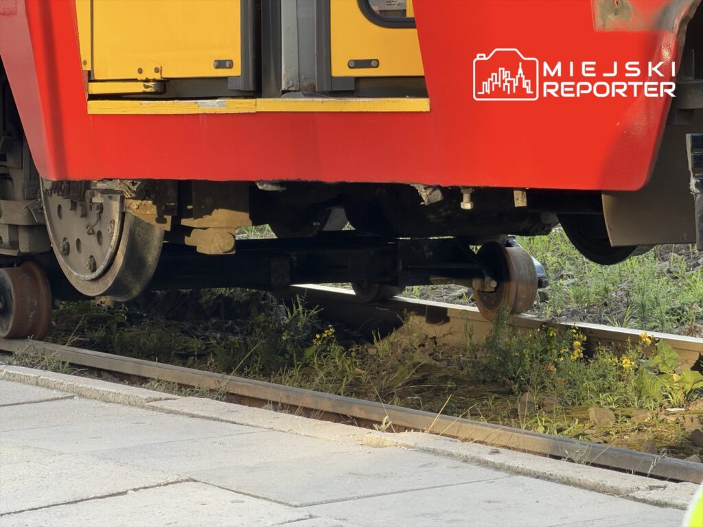 Tramwaj z czerwoną i żółtą powłoką stoi na torowisku, z jedną osią wykolejoną, otoczony trawą i kwiatami.
