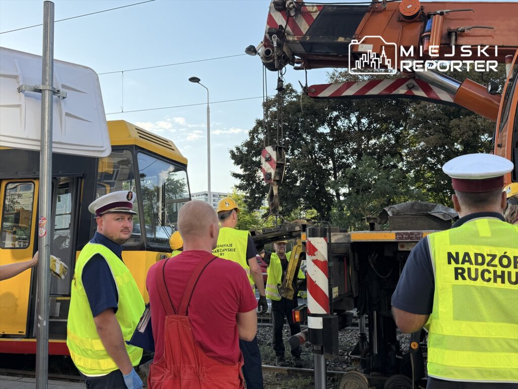 Grupa pracowników w odblaskowych kamizelkach oraz mundurach obserwuje wykolejony tramwaj, przy którym pracuje dźwig.